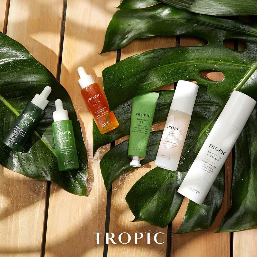 Five TROPIC skincare products arranged on large green monstera leaves over a light wood plank background. The set includes dark green, bright green, orange, and white tubes and dropper bottles, with the TROPIC brand name visible on the products and at the bottom of the image.