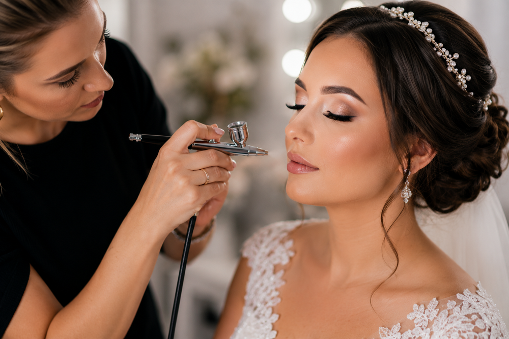 A bride in a white lace wedding gown receives makeup application from a makeup artist using an airbrush. The bride wears a pearl headpiece and veil, with a softly blurred background featuring warm lights.