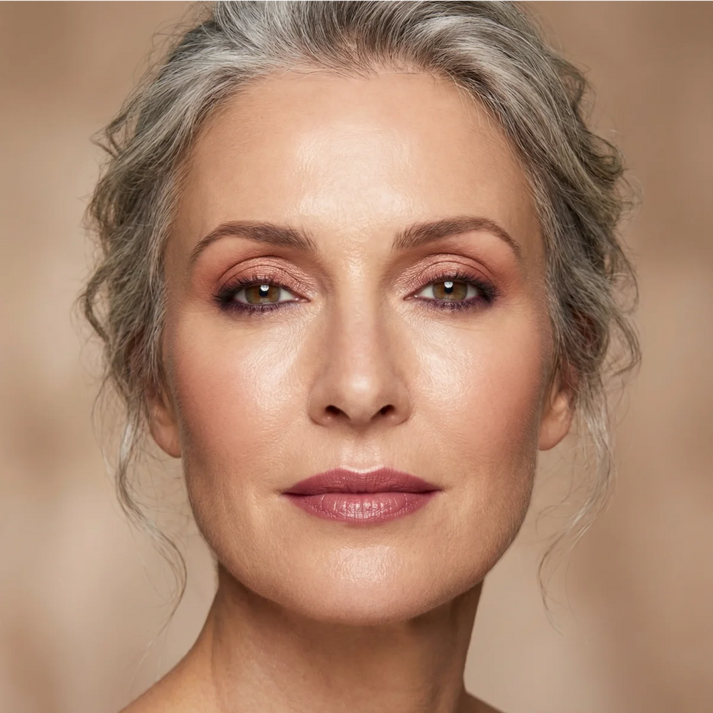 A close-up portrait of a woman with gray hair pulled back, wearing a muted mauve-pink lipstick against a soft beige background.