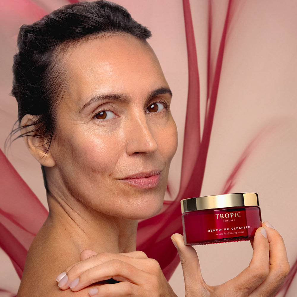 A woman holds a jar of Tropic Skincare Renewing Cleanser against a soft pink background with abstract red veining.