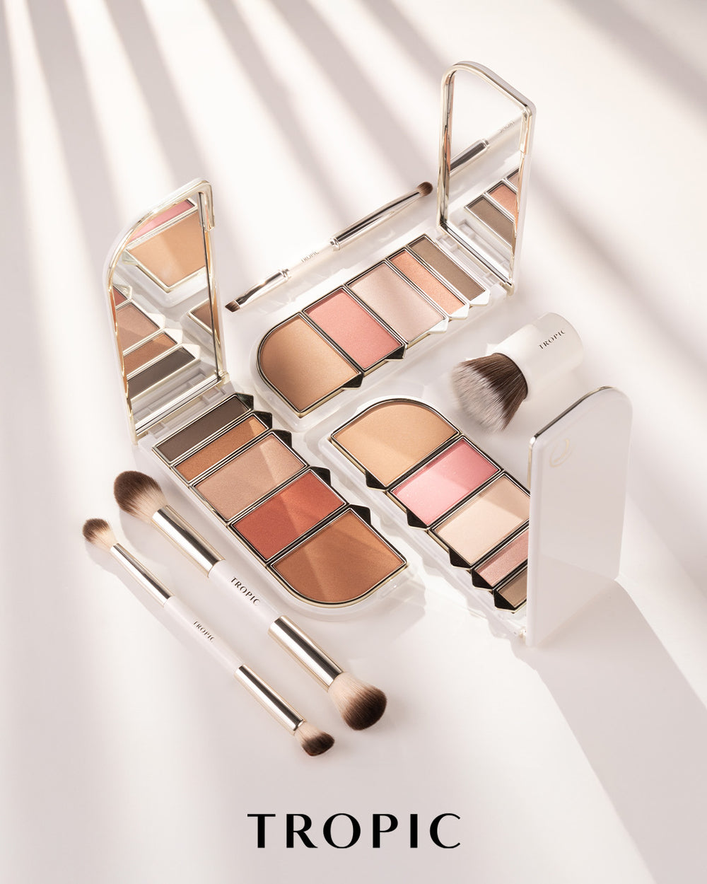 Tropic makeup palette set with multiple open eyeshadow palettes, brushes, and a stand on a white surface with diagonal shadows. The word TROPIC appears at the bottom.