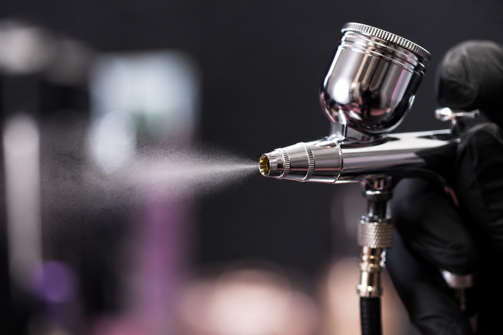 A close-up of a metallic spray gun with a black-gloved hand holding it, spraying a fine mist against a dark blurred background.