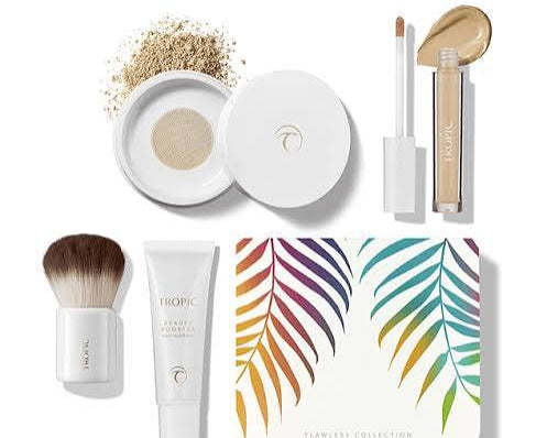 Tropic Beauty Flawless Collection makeup set on a white background, featuring a white makeup compact with gold powder, a liquid cream product, a brush, a tube, and a box with colorful palm leaf artwork.